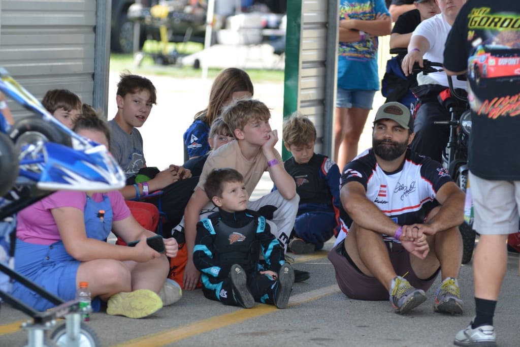 A mentor sits on the pit lane floor among young drivers during a briefing.