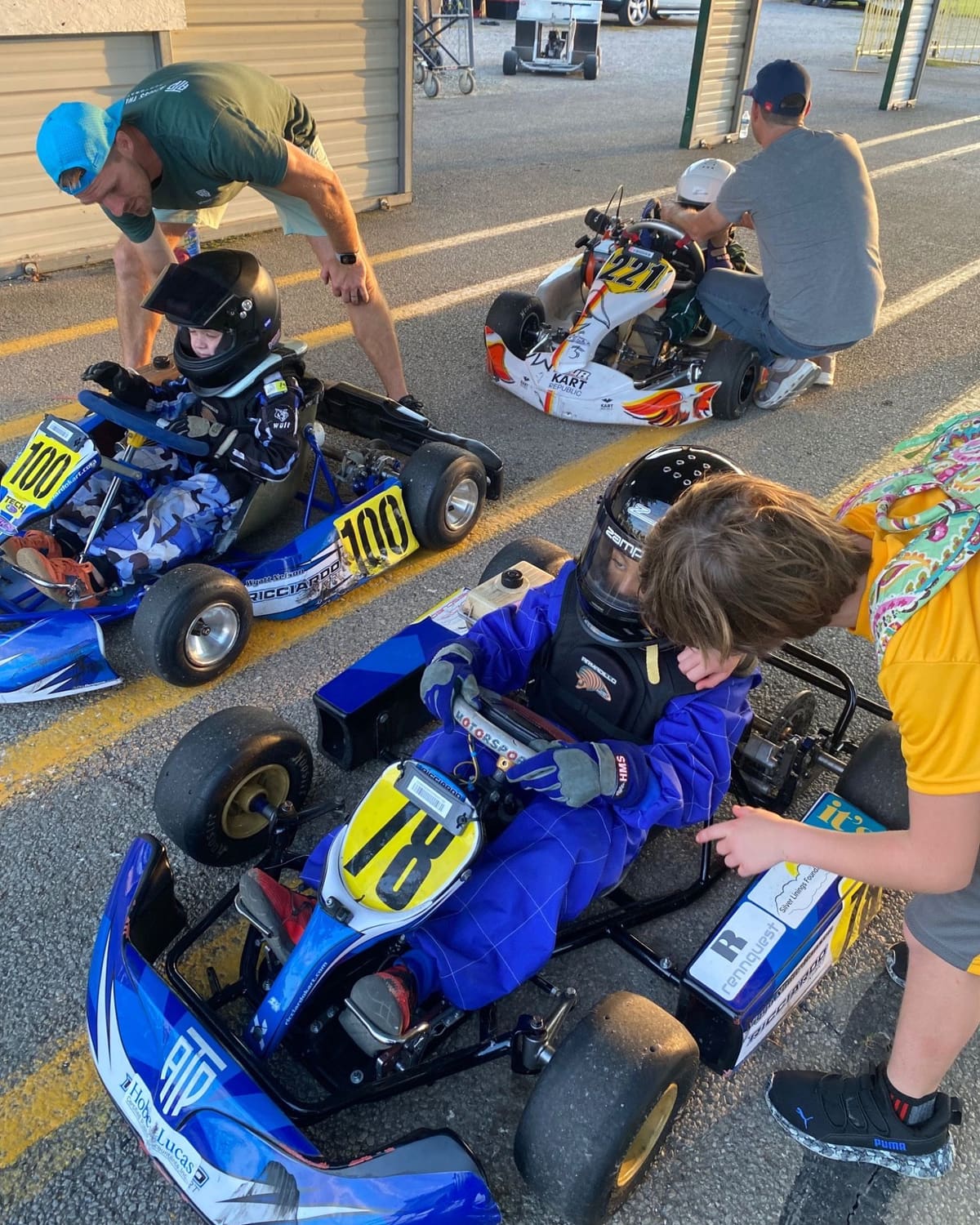 Mentors lean in to help young drivers strapped into their karts on the grid.